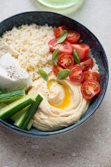 Hummus bowl with couscous, feta and fresh vegetables, vertical shot on a beige stone background, middle closeup