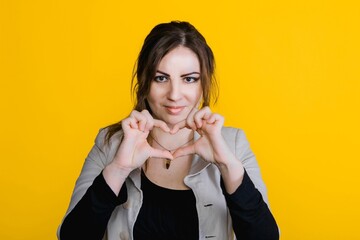 Portrait of young brunette woman in studio with yellow isolated background. Girl shows heart with...