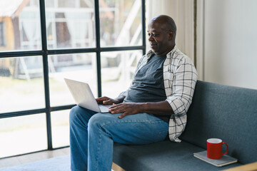 Senior Man Using Laptop at Home, Relaxing on Sofa with Coffee Mug, Casual Clothing, Natural Light, Modern Home Interior, Working Remotely, Technology and Lifestyle Concept