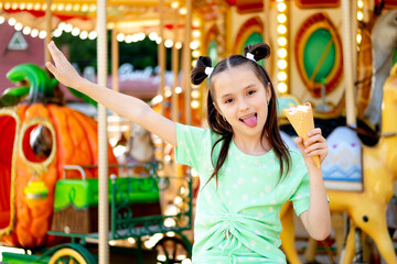 a child girl in the summer in an amusement and amusement park eats ice cream and shows her tongue and rejoices at the International Children's Day, happy childhood and holidays.