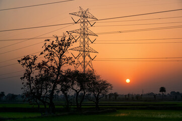 Silhouette of transmission towers carrying high voltage electricity across agricultural fields, against backdrop of setting sun in dusk. Photographed in Baghmundi, Purulia, West Bengal.