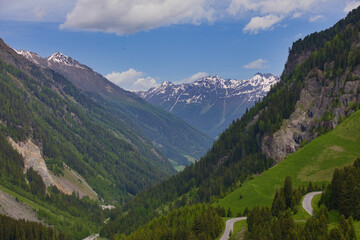 Ein atemberaubendes Bergpanorama in den österreichischen Alpen mit schneebedeckten Gipfeln, einem grünen Tal, kurvigen Straßen und Wanderwegen unter einem blauen Himmel mit weißen Wolken