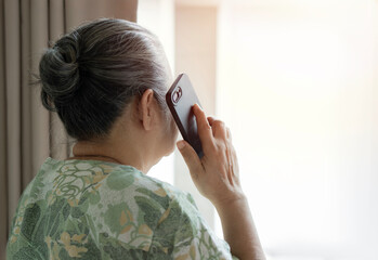 Grandma standing nearby the window and talking to family on the phone at bedroom in the morning.