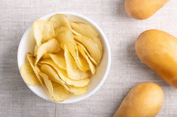 Salted potato chips in a white bowl on linen fabric. Crisps, slim slices of deep fried potato, and on the right raw potatoes, tubers of the root vegetable Solanum tuberosum. Close-up from above. Photo