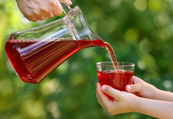 child hands holding glass and cherry juice pouring from jug with green garden on the background