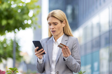 Fototapeta premium Worried businesswoman standing outdoors, holding a smartphone and credit card, expressing concern and anxiety about financial fraud or online banking issues.
