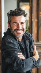 Obraz premium The image captures a cheerful, bearded man with dark, wavy hair and a noticeable gray streak. He is wearing a casual, dark-colored jacket with zippers, has his arms crossed, and is leaning comfortably