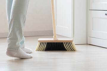 Woman using broom and sweeping light laminate floor beside white wooden furniture at home room....