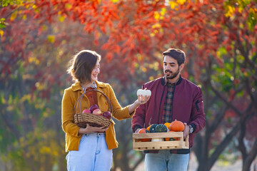 Happy caucasian farmer couple carrying organics homegrown produce harvest with apple, squash and pumpkin while walking along the country road with fall color from maple tree during autumn season