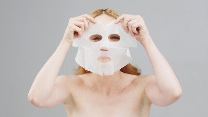 Cute, positive blonde woman applying a cosmetic fabric face mask on a white background. Daily self-care. Dermatology and skin care.
