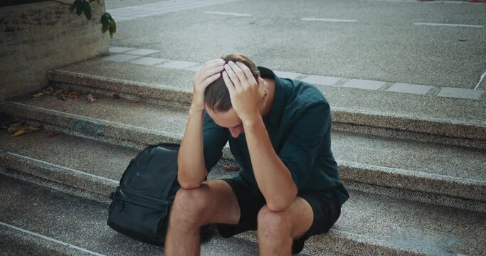 Seated urban steps caucasian mid adult man expresses stress sadness hands reaching towards head overwhelmed emotional stress. Person sits concrete steps stress sorrow turmoil emotional distress.