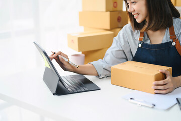 Young Woman Working on Laptop While Holding a Package, Small Business Owner, E-commerce, Online Shopping, Entrepreneur, Home Office, Shipping, Delivery, Startup, Modern Technology, Smiling