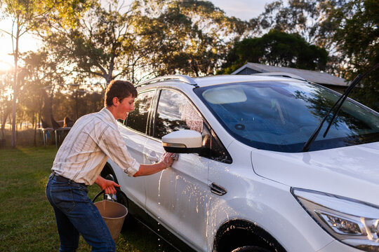 Teenager working hard washing family car in sunset light with bucket of soapy water