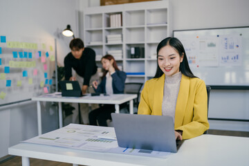 Young Professional Woman Working on Laptop in Modern Office with Colleagues Collaborating in Background, Business Charts and Sticky Notes on Walls, Bright and Productive Work Environment