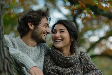 Fototapeta premium A happy couple, dressed in cozy sweaters, smiles at each other while sitting in a tree, surrounded by autumn foliage.