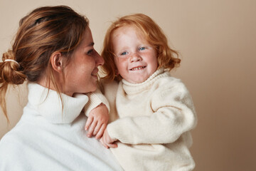 Woman holding redhaired girl in white sweater on beige background in family portrait concept