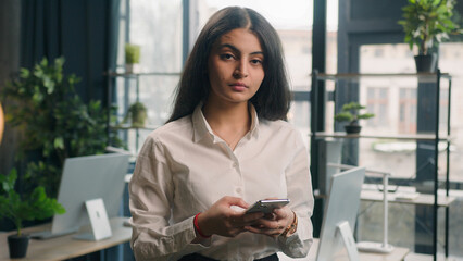 Happy multiracial woman using mobile phone in office smiling looking at camera Businesswoman Indian girl Arabian female executive worker smartphone internet surfing browsing online checking news smile