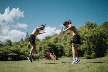 Two athletic men are performing exercises together outdoors in a park. The sun is shining, and they are surrounded by lush greenery.