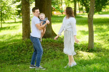 Fototapeta premium Joyful family plays with their baby in park on sunny day. Father holds girl in his arms and spins with her. Family weekend concept, happy moments