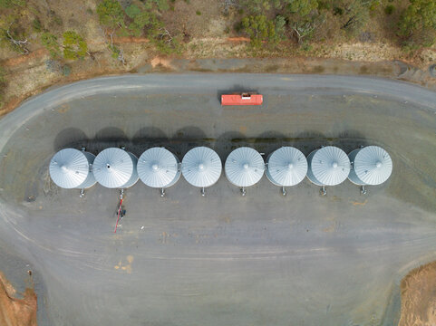 Aerial view of a line of grain storage bins in a clearing on the edge of a paddock