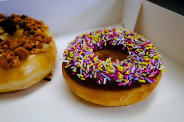 Close-up of a colorful sprinkle donut and another donut with crumble topping in a white box.