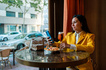 Trendy latin woman using mobile while having a beer in modern bar.