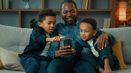 African American happy family father with ethnic sons boys little children siblings kids at home couch sofa using mobile phone together watching funny video looking at smartphone app video call chat