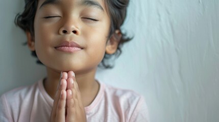 Young child with eyes closed hands clasped in prayer against a soft-focus background conveying a sense of peace and innocence.