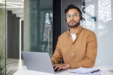 Portrait of a serious Indian man wearing a headset working in the office with a laptop, looking at...