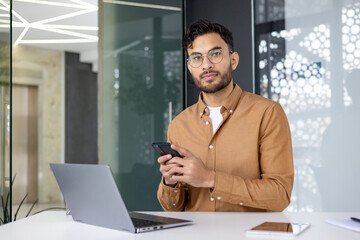 Portrait of a young Indian man in a brown shirt and glasses sitting at a desk in the office, holding a phone and looking at the camera