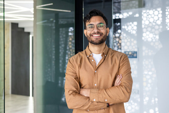 Portrait of a young Indian man wearing glasses and a brown shirt standing in a modern office, crossing his arms over his chest and looking confidently at the camera