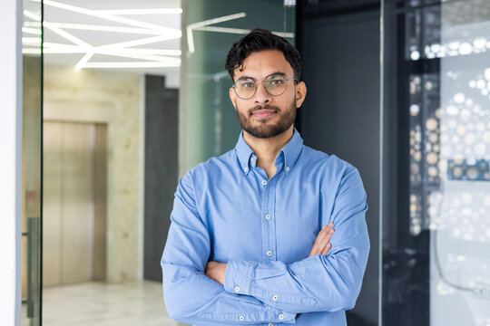 A serious Indian man in glasses and a blue shirt standing with crossed arms in a modern office and looking confidently into the camera