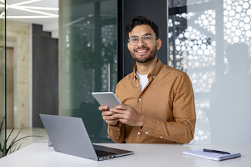 Portrait of a young Muslim man wearing glasses sitting at an office desk with a laptop, holding and...