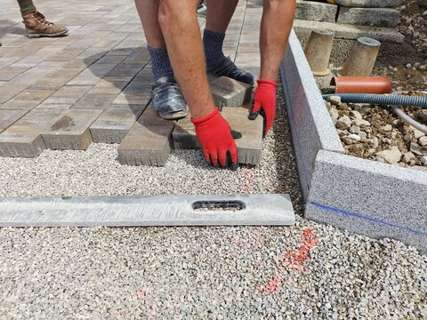 Unrecognizable male craftsman paving a forecourt with red gloves. Spirit level in gravel on the ground, ready for use. Manual work at construction site. Macro shot of paving a forecourt.