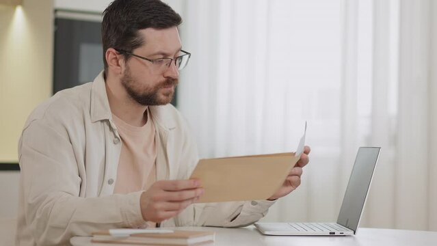 Successful caucasian male open envelope letter feeling happy to read positive news at home workplace. Bearded man in glasses expressing happy emotions on background of kitchen.