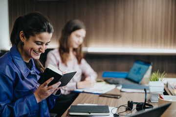 In a well-lit office, a smiling woman reads a book, enjoying a light moment, while her colleague focuses on a laptop, highlighting a pleasant, cooperative workspace atmosphere.