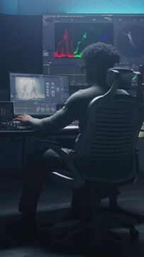 Vertical shot of African American colorist doing color grading on computer in modern studio. Multiple big monitors with action movie footage and program interface on the wall. Video post production.