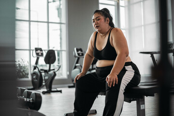 Fototapeta premium Plus size Asian woman sitting on a bench in a gym, resting after an intense workout, reflecting dedication and physical exertion.