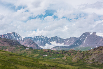 Fototapeta premium Scenic alpine landscape with green hills and rocks with view to big glacier and large snow-capped mountain range far away under clouds in blue sky in changeable weather. Awesome high snowy mountains.