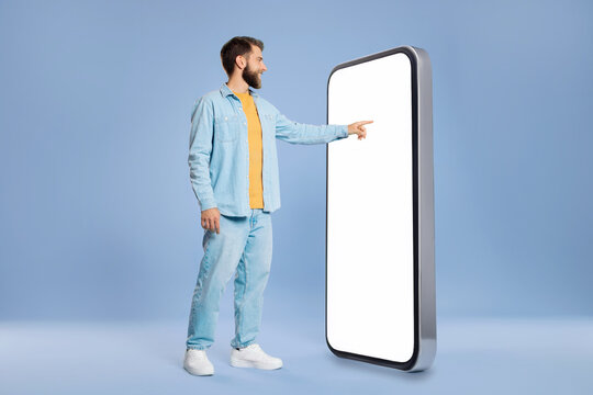 Stylish European guy touching huge smartphone blank screen, standing over blue studio background, checking newest mobile app, copy space, mockup