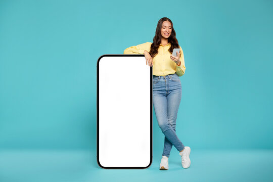 Pretty European woman posing by huge cellphone with white blank screen over blue background and using smartphone, mockup for advertisement