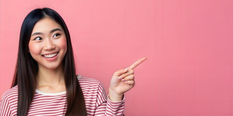 Studio shot of a positive Asian woman with a radiant smile pointing at empty space against a pink background