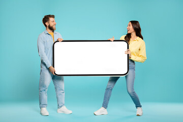 Happy young European couple fighting over big smartphone with blank white screen on blue studio background, mockup for app, ad or website