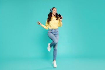 Joyful European lady in headphones singing favorite song and using cellphone as mic, dancing and jumping on turquoise background, full length