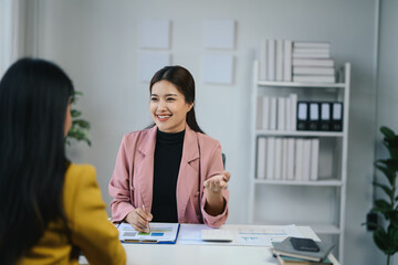 Professional Business Meeting in Modern Office with Two Women Discussing Documents and Ideas, Smiling and Engaged in Conversation, Bright and Clean Workspace with Shelves and Plants in Background