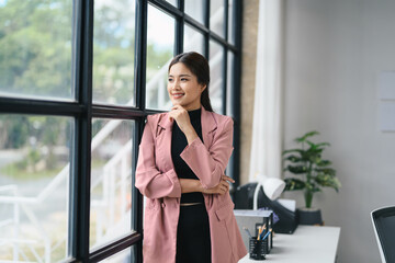 Confident Businesswoman in Pink Blazer Standing by Office Window, Smiling and Looking Outside, Modern Workspace with Natural Light and Greenery in Background