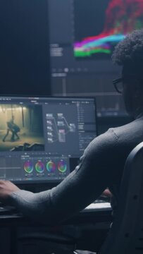 African American film editor does color grading on computer in modern studio. Big monitors with RGB colour correction graphic bar and program interface on the wall. Video post production. Vertical