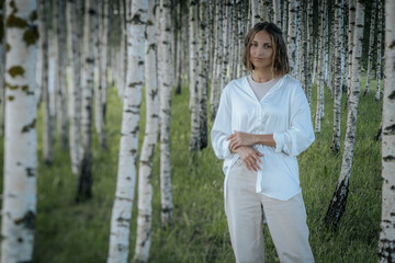 A woman with short hair and a calm expression stands in a dense birch tree forest, wearing a white shirt. The blurred background emphasizes the serene natural environment.