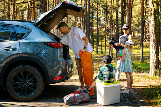 family with kids remove the camping supplies from car trunk at forest. summer vacation, road trip