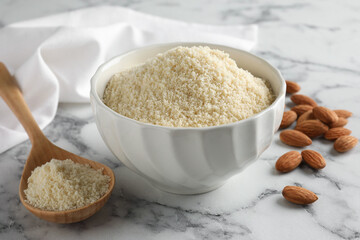 Fresh almond flour in bowl, spoon and nuts on white marble table, closeup